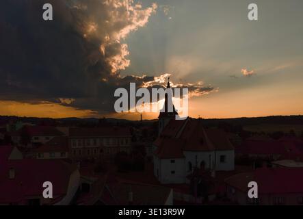 Die Kirche des Heiligen Wenzels in Netolice, die sich vor dem dramatischen Sonnenuntergangshimmel mit Sonnenstrahlen und Wolken über den historischen Dächern in Südböhmen, Tschechische Republik bildet Stockfoto