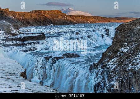 Gullfoss Wasserfall im Winter mit Eisformationen und Mt. Bláfell im Hintergrund, Island. Der teilweise gefrorene Fluss Hvitá fällt in eine tiefe Schlucht. Stockfoto
