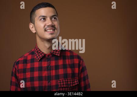 Studio shot Junger multi-ethnischen asiatischer Mann tragen rot kariertem Hemd gegen braunen Hintergrund Stockfoto