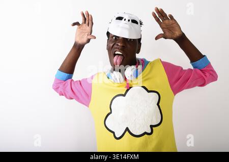 Studio shot von jungen schwarzen afrikanischen Mann die Zunge heraus, während das tragen weiße Maske auf dem Kopf und Kopfhörer um den Hals mit beiden Armen angehoben ag Stockfoto
