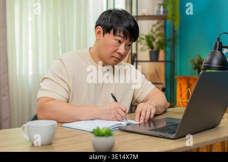 Asiatischer Mann, der zu Hause am Tisch sitzt, nimmt an einem Online-Kurs auf Laptop Teil, schreibt Notizen und erklärt der Kamera, wie sie sich zeigt. Der Chinese spricht selbstbewusst, mit ausdrucksstarken Gesten, um ansprechende Online-Lektion zu erstellen. Stockfoto