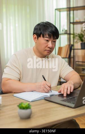 Asiatischer Mann, der zu Hause am Tisch sitzt, nimmt an einem Online-Kurs auf Laptop Teil, schreibt Notizen und erklärt der Kamera, wie sie sich zeigt. Der Chinese spricht selbstbewusst, mit ausdrucksstarken Gesten, um ansprechende Online-Lektion zu erstellen. Stockfoto
