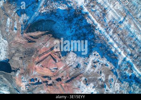 Industrieabfall zeigt Schichten roter Bergbauabfälle gegen kalten Winterschnee. Ein eindrucksvoller Abstract der Bodendegradation, der Rohstoffgewinnung und des ökologischen Fußabdrucks der Schwerindustrie in einer kargen Landschaft. Stockfoto