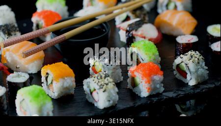 Verschiedene Arten von Sushi-Brötchen mit Stäbchen, die während der Mittagszeit auf einem schwarzen Teller an einem Esstisch platziert werden Stockfoto