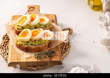 Zwei Scheiben knuspriges Sauerteigbrot, belegt mit Avocadopüree und Marmeladen-Eiern. Das helle Morgenlicht erzeugt sanfte Schatten auf dem weißen Holztisch Stockfoto