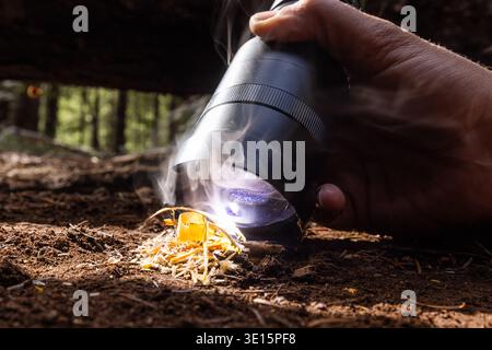 Eine leistungsstarke Taschenlampe löst ein Feuer auf, wenn sie entzündet wird. Die Finger greifen die Taschenlampe, durchgehend, fokussiert. Das Feuer knistert tief unter dem Waldboden. Dreck, Zweige und Leaf Stockfoto