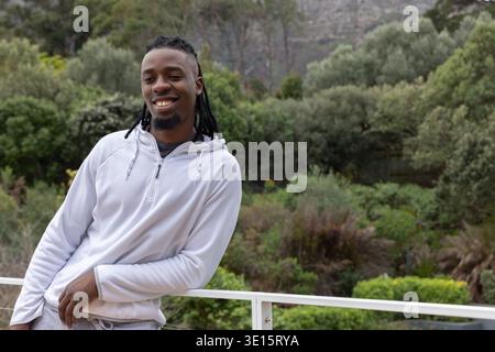 Afroamerikaner lehnt sich auf weißes Metall-Balkongeländer, lächelt mit langen Locs, trägt Hoodie Stockfoto