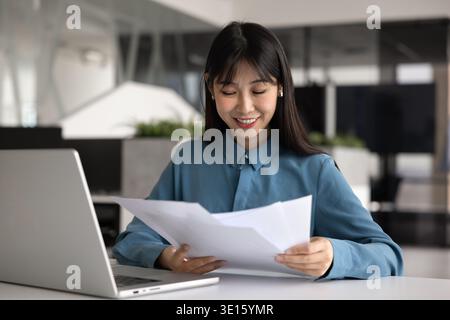 Positive junge chinesische Projektmanagerin, die Papier am Arbeitsplatz überprüft Stockfoto