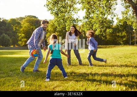 Lächelnde Eltern und Kinder spielen gemeinsam auf dem grünen Rasen Ball und genießen Outdoor-Aktivitäten im Park. Stockfoto