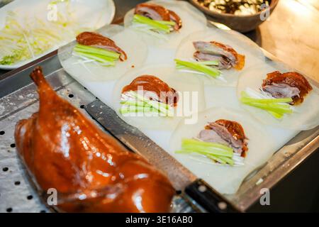 Der fachkundige Küchenchef schneidet knusprige peking-Ente mit frischen Frühlingszwiebeln, Gurkenscheiben und Hoisin-Sauce auf zarten Pfannkuchen und präsentiert sie Stockfoto