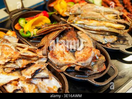 Auswahl an goldgegrillten und in der Pfanne frittierten Fischfilets mit bunten Paprika und Gemüse, heiß in gusseisernen Töpfen serviert für eine lebendige Atmosphäre Stockfoto