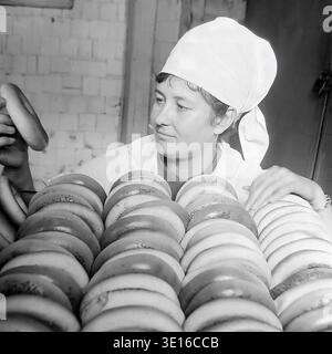 Ein faszinierendes Schwarzweißfoto aus den 1960er Jahren zeigt eine engagierte Bäckerin, die eine riesige Menge frisch gebackener Bubliki in einer Slawjansk-Brotfabrik sorgfältig inspiziert. Sie hält ein traditionelles Ringbrot hoch, während sie dicht hinter endlosen Reihen des goldenen Teigs steht. Dieses wunderschöne Vintage-Bild zeigt die authentische sowjetische Lebensmittelherstellung, Qualitätskontrollverfahren und industrielle Arbeit aus der Mitte des Jahrhunderts perfekt. Dieses Archivbild spiegelt den Alltag des friedlichen Donbass vor dem Krieg wider. Stockfoto