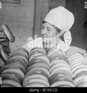 Ein faszinierendes Schwarzweißfoto aus den 1960er Jahren zeigt eine engagierte Bäckerin, die eine riesige Menge frisch gebackener Bubliki in einer Slawjansk-Brotfabrik sorgfältig inspiziert. Sie hält ein traditionelles Ringbrot hoch, während sie dicht hinter endlosen Reihen des goldenen Teigs steht. Dieses wunderschöne Vintage-Bild zeigt die authentische sowjetische Lebensmittelherstellung, Qualitätskontrollverfahren und industrielle Arbeit aus der Mitte des Jahrhunderts perfekt. Dieses Archivbild spiegelt den Alltag des friedlichen Donbass vor dem Krieg wider. Stockfoto