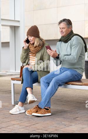 Seniorenpaar sitzt auf einer Holzbank vor dem Gebäude, eine Frau, die telefoniert, ein Mann, der das Telefon benutzt Stockfoto