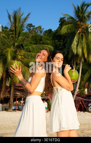 Zwei lächelnde Frauen stehen an einem tropischen Strand zurück an Rücken und halten während des Sonnenuntergangs frische Kokosgetränke Stockfoto