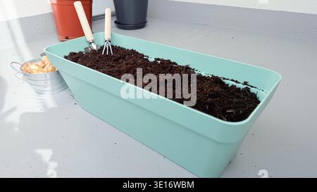Mintgrüner Pflanzgefäß gefüllt mit Boden und Gartenwerkzeugen auf einer Terrasse mit sichtbaren Topfpflanzen Stockfoto