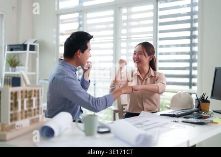 Zwei lächelnde asiatische Kollegen feierten den Projektabschluss. Architekturpläne und Schutzhelm in moderner Büroumgebung. Stockfoto
