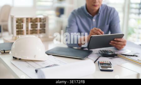 Ein Erwachsener Architekt des asiatischen Mannes prüft Baupläne mit Tablet-Technologie. Fokussierte professionelle Entwürfe für neues Gebäudeprojekt in hellen Büros. Stockfoto
