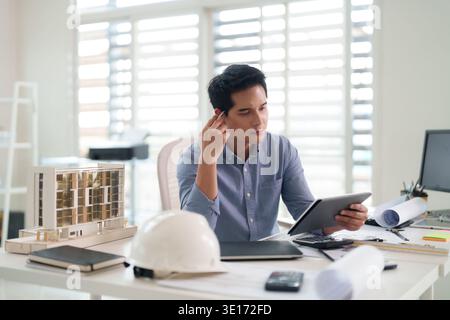 Asiatischer Architekt prüft Blueprints Digital Tablet. Professionelle, Mann fokussierte Projektentwicklung modernes Büro. Technische Innovation Gebäudedesign. Stockfoto