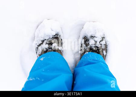 Ein winterlicher Blick von oben mit schneebedeckten Stiefeln und hellblauen Hosen auf Neuschnee Stockfoto