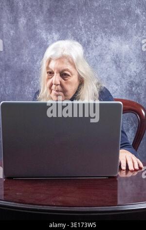 70-jährige Frau, die vor einem Laptop sitzt. Die Frau ist kaukasischer Herkunft, hat lange blonde Haare und trägt ein Rollkragenhemd Stockfoto