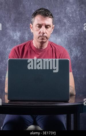 Mann, der vor einem Laptop sitzt. Der Mann ist kaukasischer Herkunft, vierzig Jahre alt und trägt ein burgunderrotes T-Shirt Stockfoto