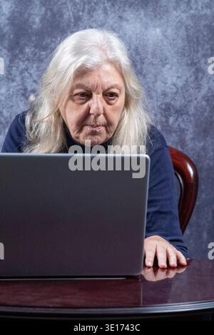 70-jährige Frau, die vor einem Laptop sitzt. Die Frau ist kaukasischer Herkunft, hat lange blonde Haare und trägt ein Rollkragenhemd Stockfoto