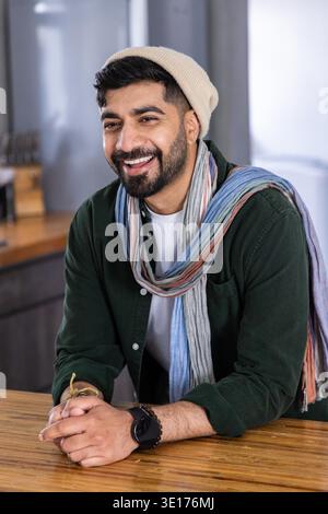Indischer Mann lehnt sich auf Holztheke in der Küche und lächelt, während er einen Mütze-Schal und Smartwatch trägt. Porträt, Bartop, gemütlich, warm, innen, modern, Lifestyle || Model veröffentlicht Stockfoto
