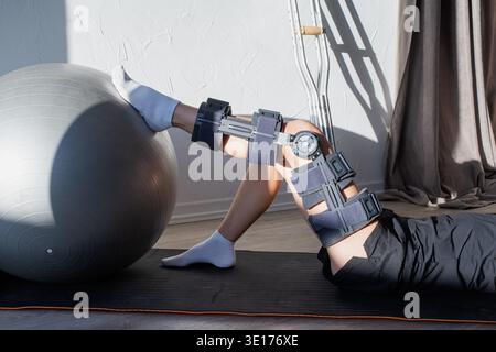 Rehabilitation nach Knieoperationen. Frau mit Sportkleidung und Orthese, die Übungen mit Fitnessball macht Stockfoto