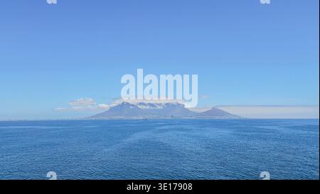 Panoramablick aus der Ferne auf Kapstadt vom Meer aus mit tiefblauem Wasser, subtilen Strömungen, Tafelberg mit Wolken und schmaler Stadtstraße an der Basis Stockfoto