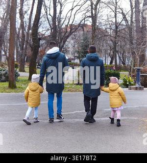Zwei Männer halten Hände mit zwei Mädchen in gelben Jacken, während sie durch einen Park laufen, umgeben von Bäumen und städtischen Gebäuden im Hintergrund Stockfoto