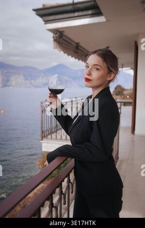Eine Frau strahlt Eleganz aus, während sie auf einem Balkon mit Blick auf das ruhige Wasser steht. In einem eleganten schwarzen Anzug hält sie ein Glas Rotwein und verkörpert Gra Stockfoto