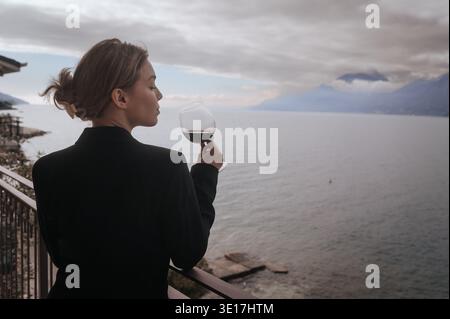 Eine Frau strahlt Eleganz aus, während sie ein Glas Rotwein auf einem Balkon am See genießt. Der ruhige Blick auf das Wasser und die Berge sorgt für eine ruhige Atmosphäre, epi Stockfoto