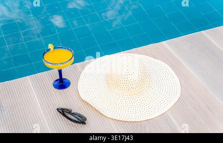 Eine ruhige Sommerszene mit einem erfrischenden Cocktail neben einem Swimmingpool, ergänzt durch einen stilvollen Sonnenhut und eine Sonnenbrille, sorgt für Entspannung und Entspannung Stockfoto