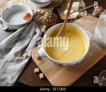Kuchenteig in Keramikschale mit Holzlöffel auf bemehltem Brett mischen. Hochwinkelschuss aus rohen gelben Teigen in einem floralen Keramiktopf, umgeben von Backofen Stockfoto