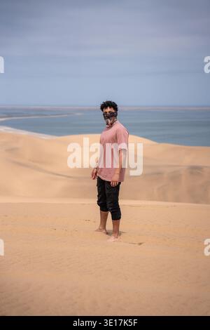 Ein Mann in lässiger Kleidung steht auf den goldenen Dünen der Walvis Bay, Namibia, mit dem Atlantischen Ozean im Hintergrund, trägt einen Schal und blickt in den Blick Stockfoto