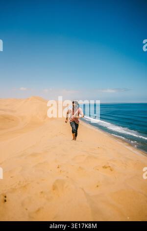 Eine Person, die eine goldene Sanddüne in Walvis Bay, Namibia, mit dem weitläufigen Ozean im Hintergrund unter einem klaren blauen Himmel hinaufläuft und Abenteuer zeigt Stockfoto
