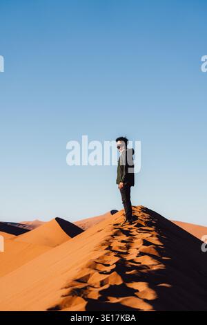 Ein Alleinreisender steht auf der Dune 45 in Namibia und bestaunt die atemberaubende Wüstenlandschaft. Die tieforange Sanddünen erstrecken sich endlos unter einem hellen B Stockfoto