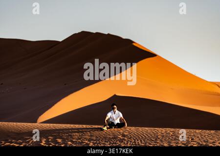 Ein Mann sitzt ruhig auf den riesigen Dünen von Sossusvlei in Namibia, beleuchtet von der goldenen Sonne. Der satte orange Sand und der klare Himmel schaffen ein atemberaubendes l Stockfoto