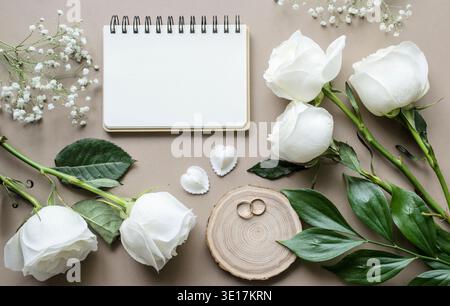 Romantisches Modell mit Blumen und einem Notizbuch auf beigefarbenem Hintergrund. Rosen und ein Notizbuch mit Platz für Text. Hochzeitskonzept. Blick von oben. Stockfoto