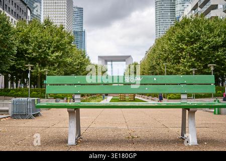 Eine pulsierende grüne Parkbank aus Holz mit Graffiti-Markierungen befindet sich im Vordergrund eines modernen städtischen plaza in Paris, Frankreich Stockfoto