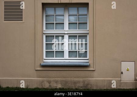 Weißes Mehrscheibenfenster an einer beigefarbenen Putzwand. Minimalistische Fassade mit einem Lüftungsgitter aus Metall und einem kleinen Schaltkasten unten Stockfoto