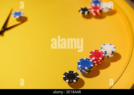 Stapel von Poker-Chips, die auf einem hellgelben Tisch in der Nähe der Uhrzeiger angeordnet sind, deuten auf Zeitdruck im Casino-Glücksspiel-Konzept, Glücksspiel, Casino-Enterta hin Stockfoto