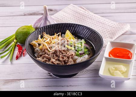 Aromatische Schüssel mit traditionellem vietnamesischem Pho Bo mit Rindfleisch, Nudeln und Sojasprossen. Serviert mit Limette, Chili und Knoblauch auf weißem Holzhintergrund. F Stockfoto