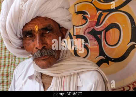 Ein Porträt zeigt einen älteren Mann in Puschkar, der einen großen weißen Turban und einen tilak auf der Stirn trägt und vor einem gemalten OM-Symbol posiert. Stockfoto