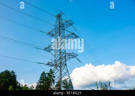 Ein markanter Power-Pylon aus Metall dominiert den Rahmen, der Stromleitungen gegen einen leuchtend blauen Himmel mit verstreuten Wolken und entferntem grünen Laub trägt Stockfoto