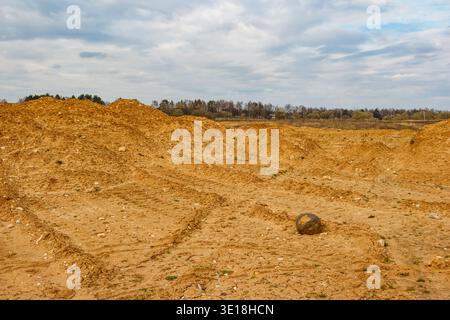 Abgerundeter Felsbrocken liegen deutlich auf kargem Steinbruchgrund unter einem bewölkten Frühlingshimmel. Ferne Erdhügel markieren das verwüstete Land Stockfoto