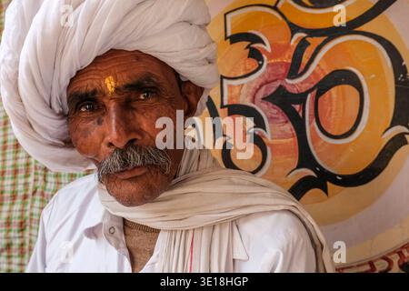 Ein Porträt zeigt einen älteren Mann in Puschkar, der einen großen weißen Turban und einen tilak auf der Stirn trägt und vor einem gemalten OM-Symbol posiert. Puschkar Ra Stockfoto