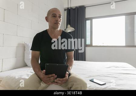 Hispanischer reifer Glatzkopf in den 40er Jahren, der auf dem Bett sitzt, während er einen digitalen Tablet-Computer in seiner modernen Wohnung benutzte, aufgenommen mit Weitwinkelobjektiv Stockfoto