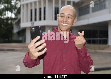 Glatzkopf italienischer Mann in burgunderrotem Hemd, der sein Smartphone überprüft, während er vor dem Universitätsgebäude bei hellem Sonnenlicht steht. Das Konzept des digitalen Kombi Stockfoto